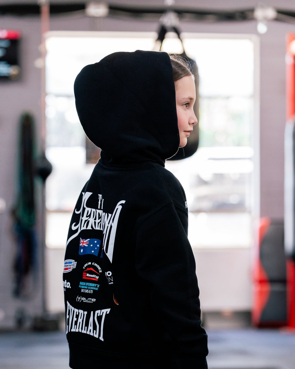 Side view of a girl in the kids black Team Teremoana Hoodie with the hood up, showing the back print