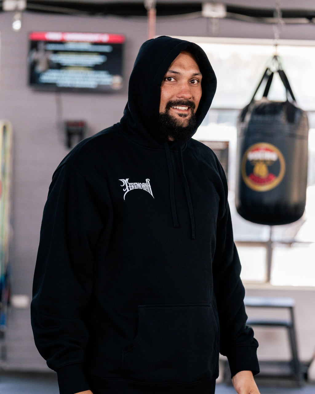 Teremoana in the black Team Teremoana Hoodie with the hood up, smiling in the gym