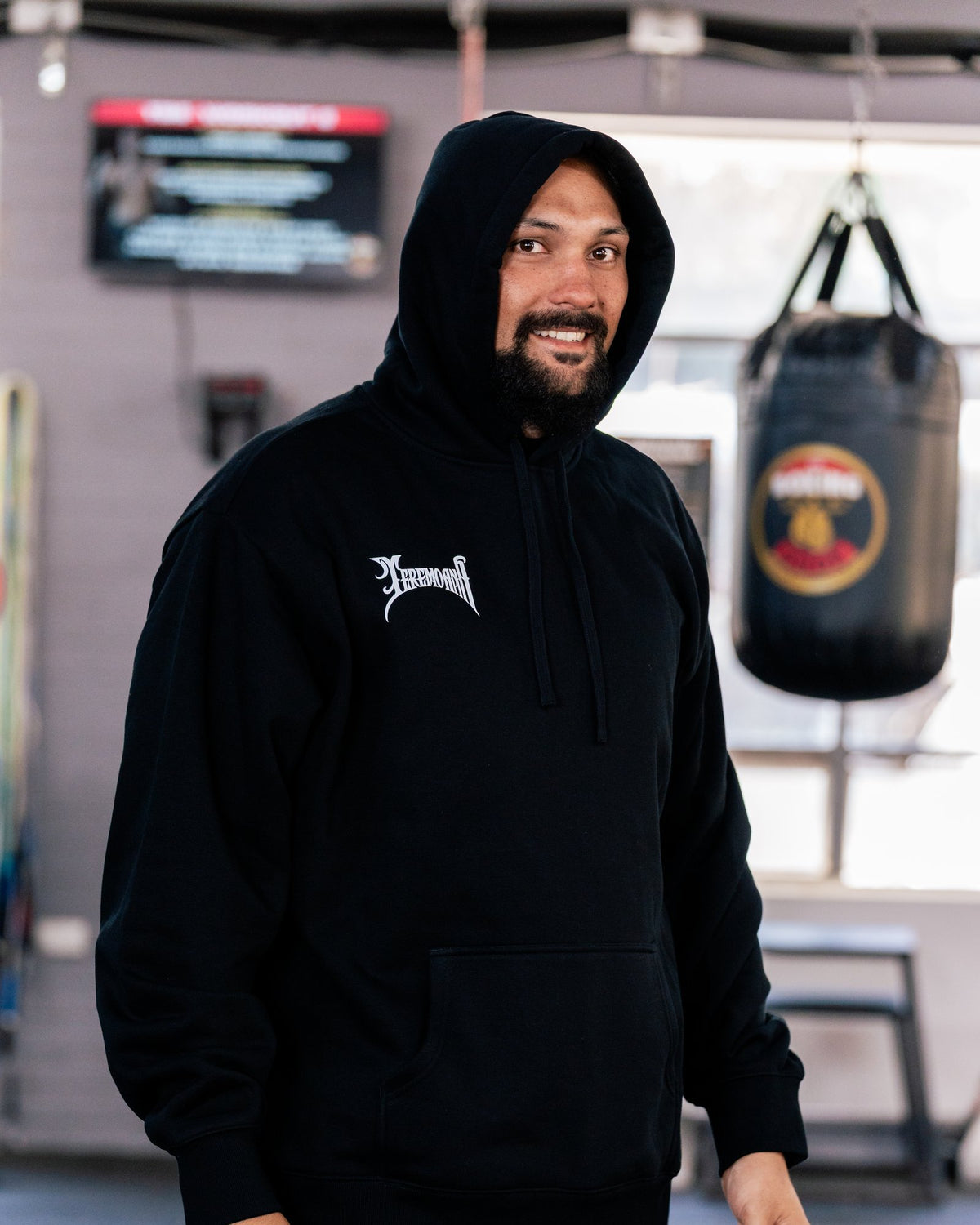 Teremoana in the black Team Teremoana Hoodie with the hood up, smiling in the gym